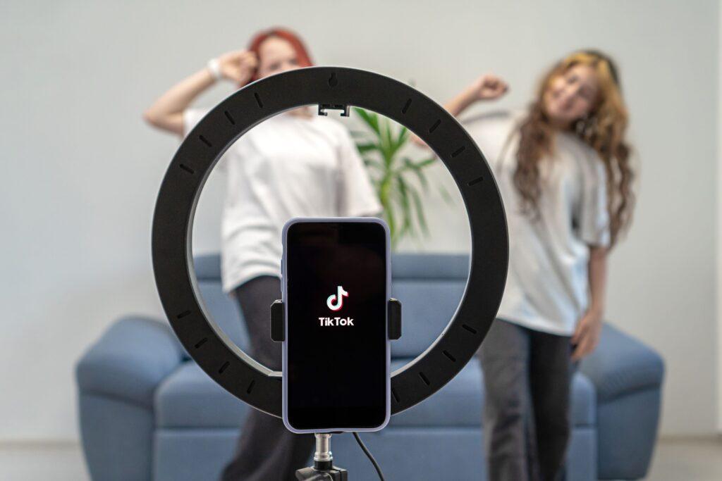 Zwei Mädchen im Teenageralter drehen bei sich zu Hause ein Video für ihren Tiktok-Vlog. Tiktok-Logo auf einem Telefon auf einem Stativ. Talente der Generation Z spielen eine Video-Selfie-Shooting-App, um eine virale Story zu teilen.