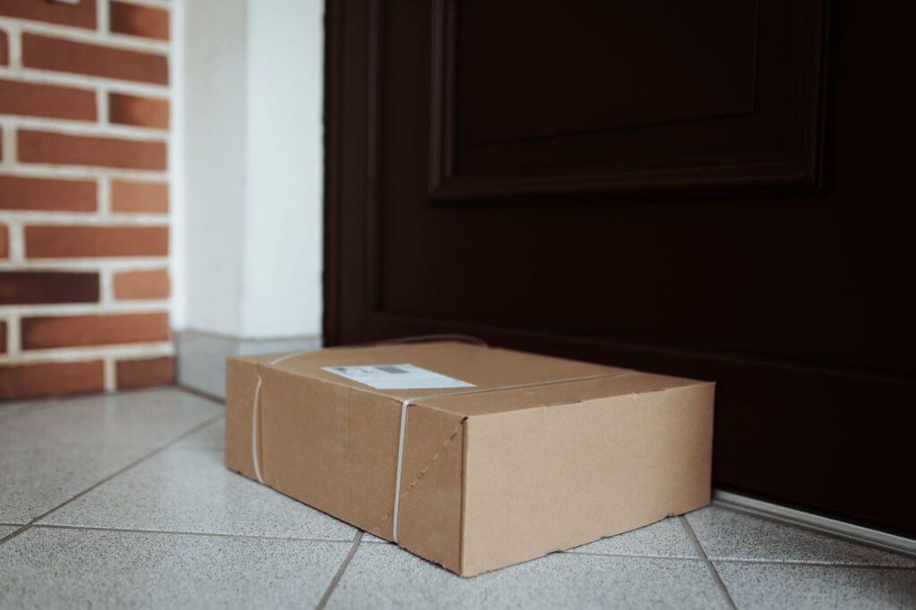 Ein ungesichertes Paket auf einer gefliesten Veranda nahe einer dunkelbraunen Tür, das die Notwendigkeit eines sicheren Paketkastens zur Verhinderung von Diebstahl und Sicherstellung der Paketsicherheit hervorhebt.