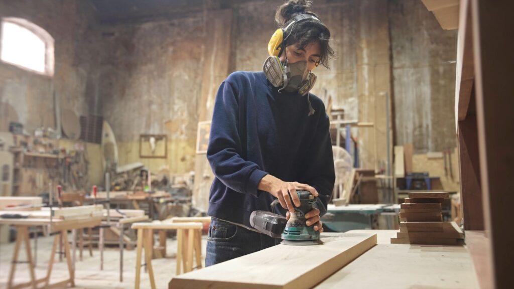 Handwerkerin beim Schleifen von Holz in einer Werkstatt, um einen maßgefertigten Couchtisch zu fertigen, mit Fokus auf handwerkliche Präzision und hochwertige Materialien.