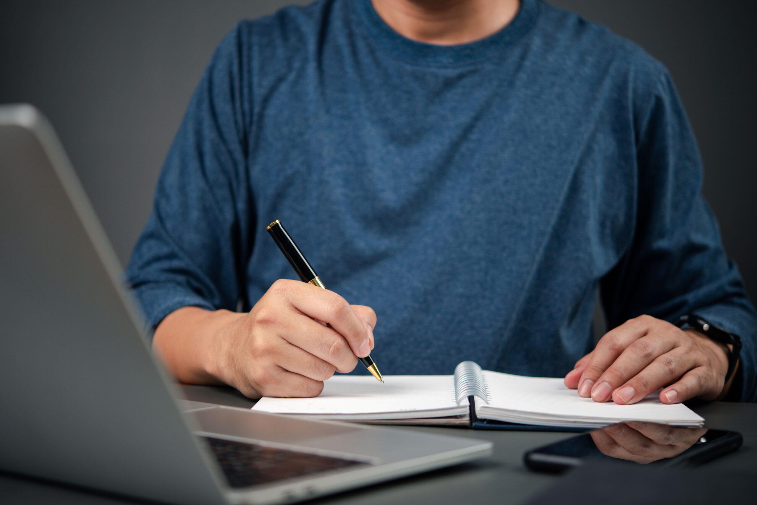 Person schreibt mit Stift in Notizbuch vor Laptop – Symbolbild für improvisierte E-Learning Produktion