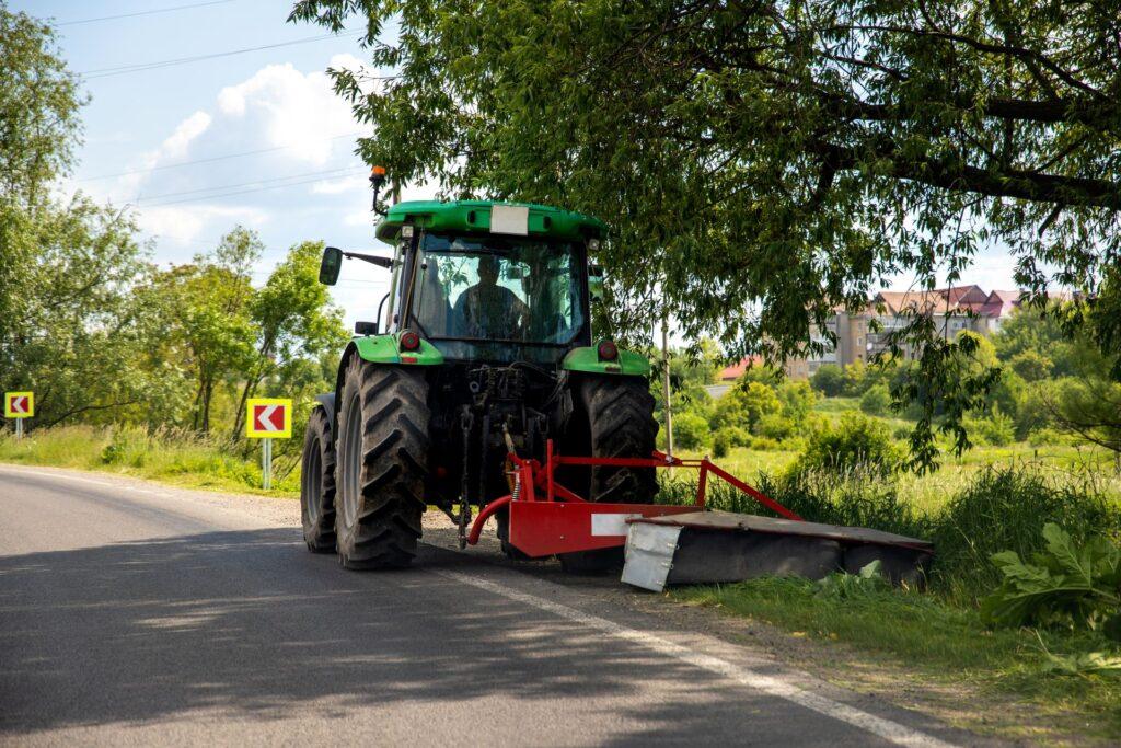 Traktor mit rotem Seitenanbau bei Maeharbeiten am Rand einer Landstrasse in gruener Umgebung